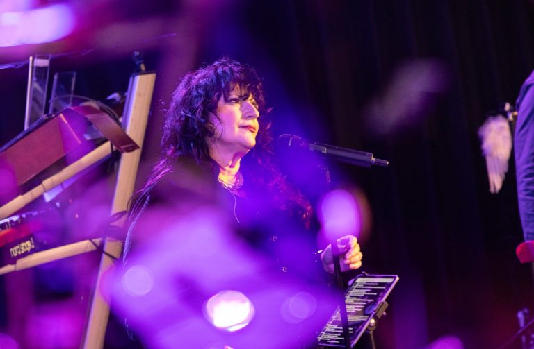 Frau mit Locken singt am Mikrofon, umgeben von buntem Licht.